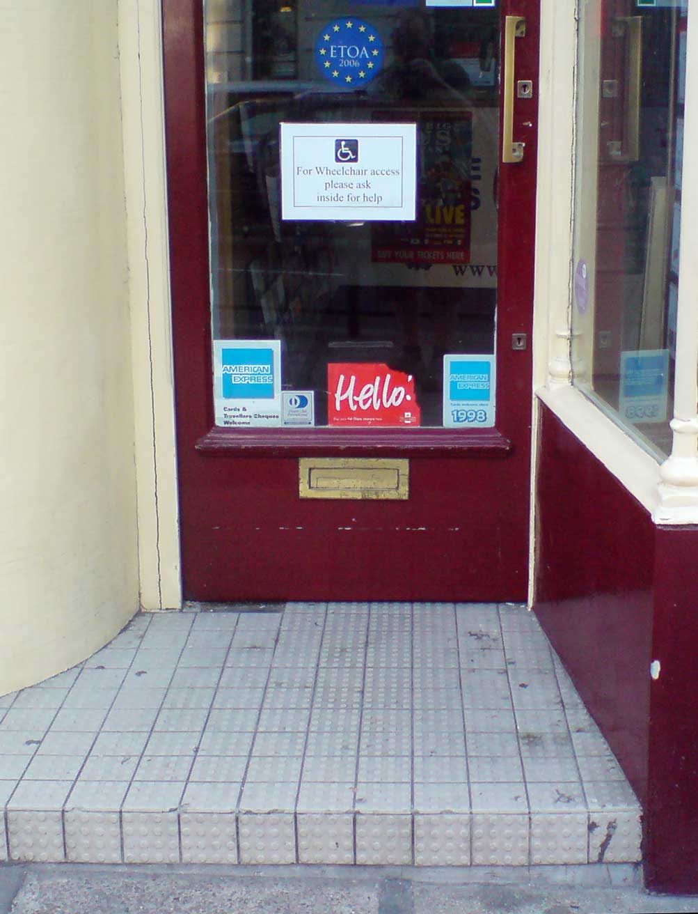 Door with a sign, “For Wheelchair access please ask inside for help.” In front of the door is a roughly 4-inch high step. Image by Darren Foreman.
