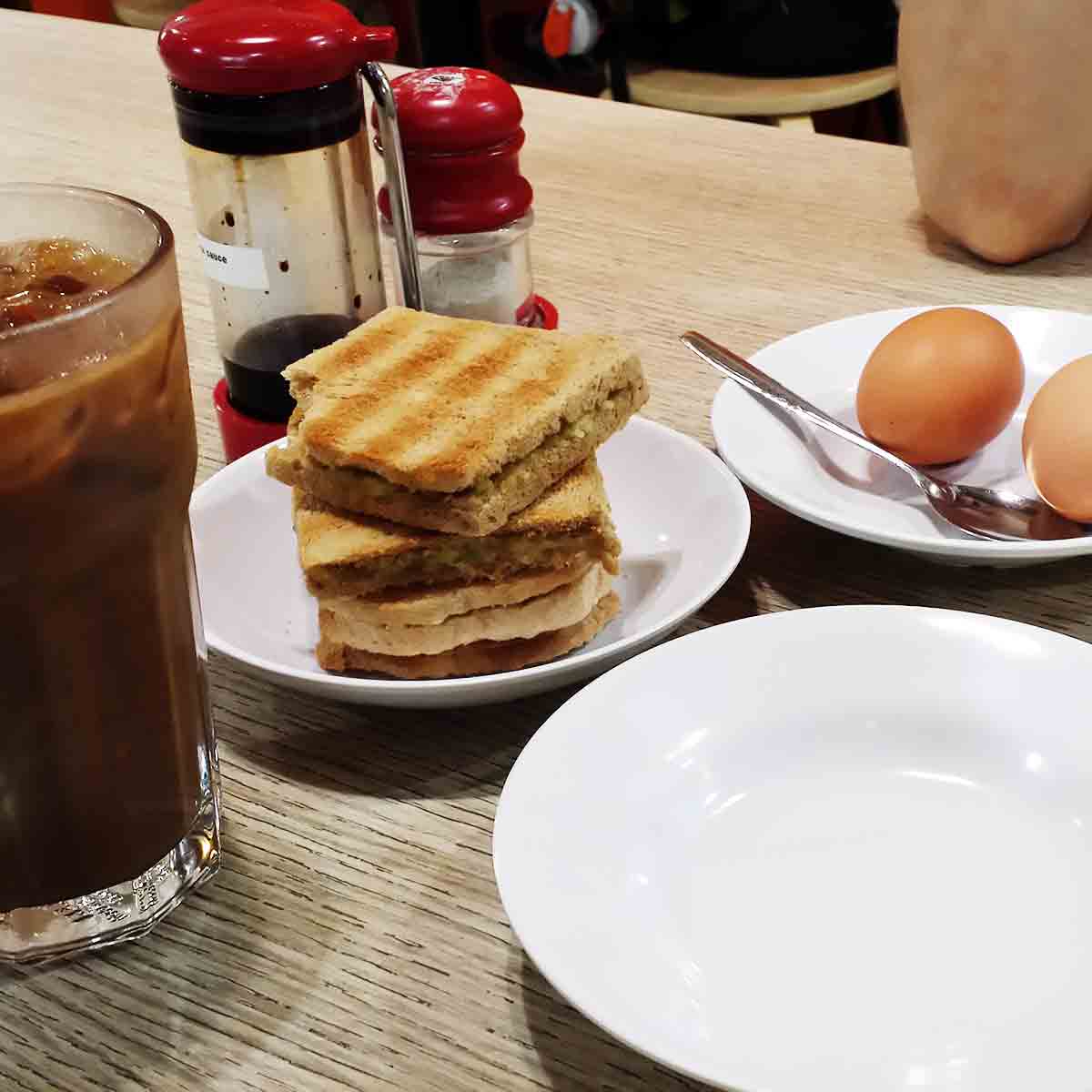 Glass of iced coffee, a stack of squares of kaya toast, a plate with soft-boiled eggs in the shell, a caddy with white pepper and soy sauce containers.