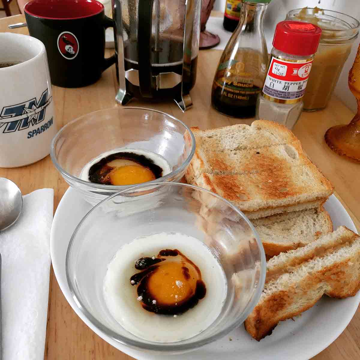 One poached egg each in two custard cups, with a bit of soy and white pepper. The cups are on a plate alongside a stack of kaya toast (kaya and butter sandwiched between toast cut into strips). In the background is a coffee mug next to a French press, along with the condiments used in the breakfast.