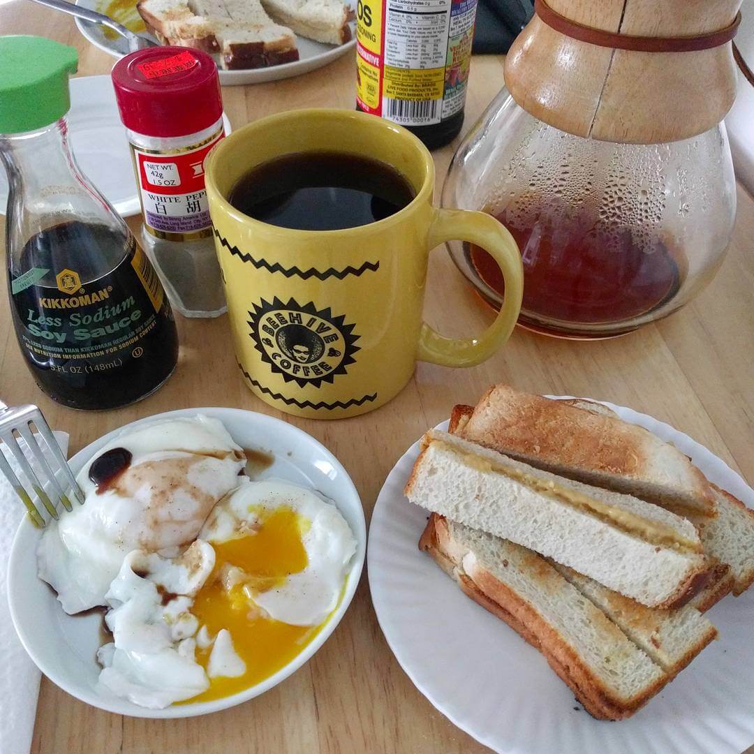 Poached eggs in a saucer with soy sauce and white pepper, one egg is broken so the yolk is flowing. Another plate with strips of kaya toast (toasted country white with kaya jam between slices, cut into strips). A yellow coffee mug with the Beehive Coffee logo full of coffee. A Chemex, some low-sodium soy sauce, and white pepper are visible in the background.