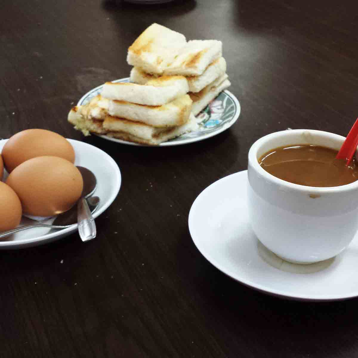 A plat of kaya toast strips, a plate with five soft-boiled eggs, a saucer and cup with coffee and cream and an odd red plastic spoon.