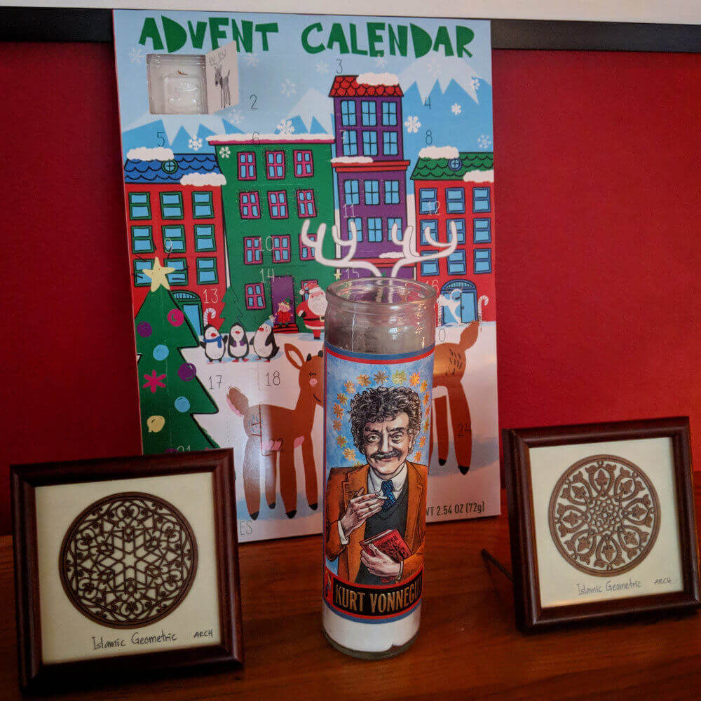 A cardboard advent calendar behind a Kurt Vonnegut saint candle and two framed Islamic geometric design prints.