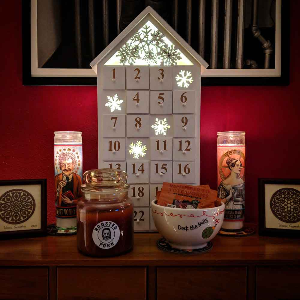 A wooden advent calendar flanked by two tall candles (one of Ada Lovelace, one of Vonnegut), and two Islamic geometric woodcuts. In the foreground is a large brown candle labeled ‘roasted pork’ and a bowl of gummi worms. All this on a wooden card catalog against a red wall.