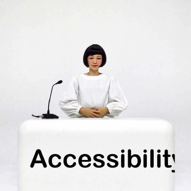A female-appearing android dressed in white sits behind a white desk in a white room. Across the front of the desk is the word ‘Accessibility’, except it does not fit on the desk.