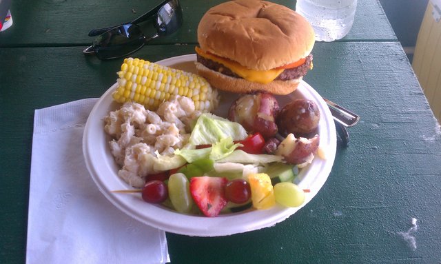 First plate: corn on the cob, salad, cheese burger, macaroni salad, fruit skewers, red potatoes.