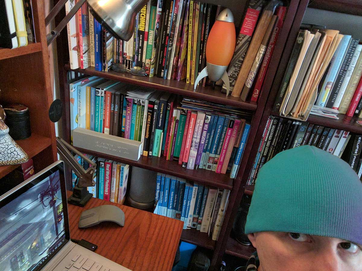 Me wearing a teal beanie with much of my bookshelf visible in the background.