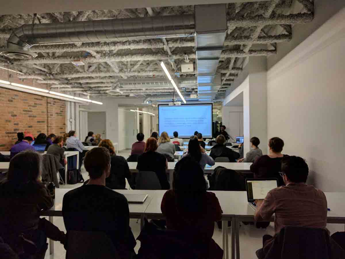 Photo showing much of the length of the room at HackerYou with many people at tables watching the slides.