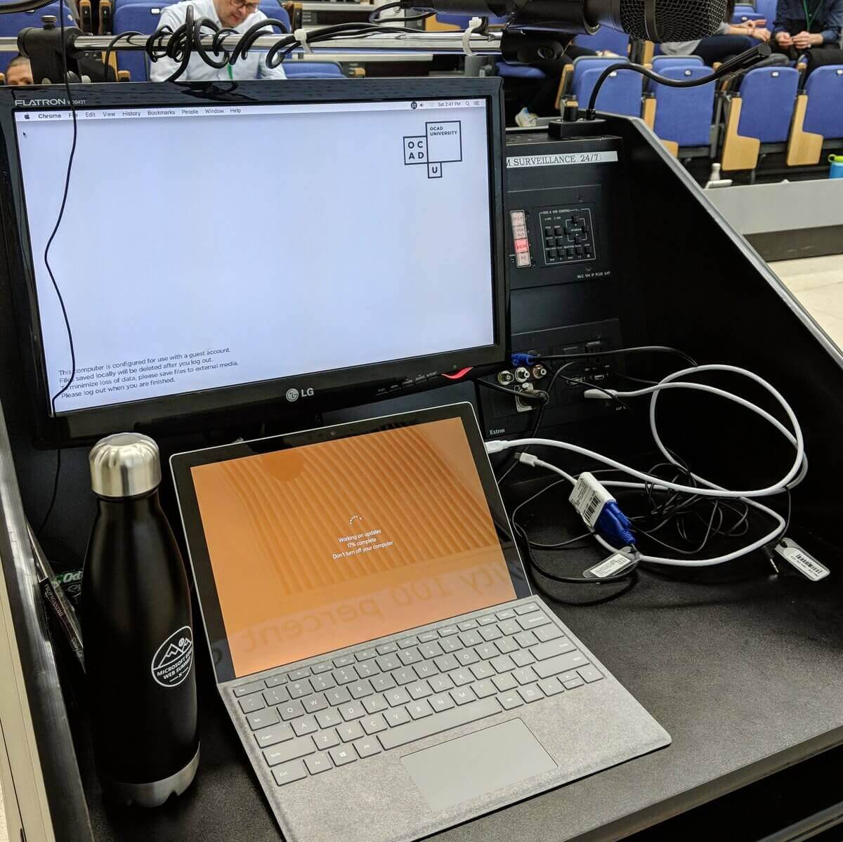 A laptop on a lectern with a gathering crowd in the seats in the background. The laptop screen says “Working on updates. 17% complete. Don’t turn off your computer.”