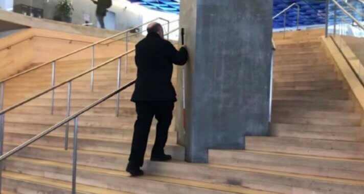 a blind man collides with a large pillar that interrupts a handrail going up the middle of a large staircase