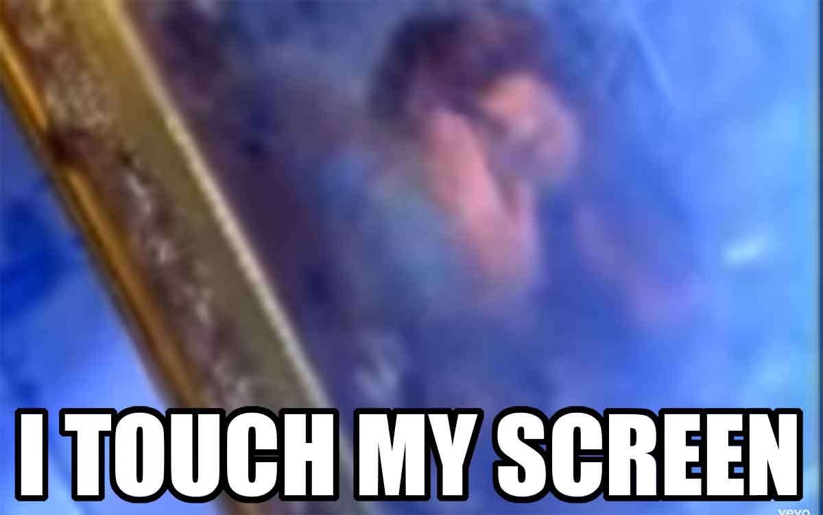 A woman holding her hand up to her reflection in a mirror, all within a smokey blue-lit scene from a music video, overlaid with the text ‘I touch my screen’.