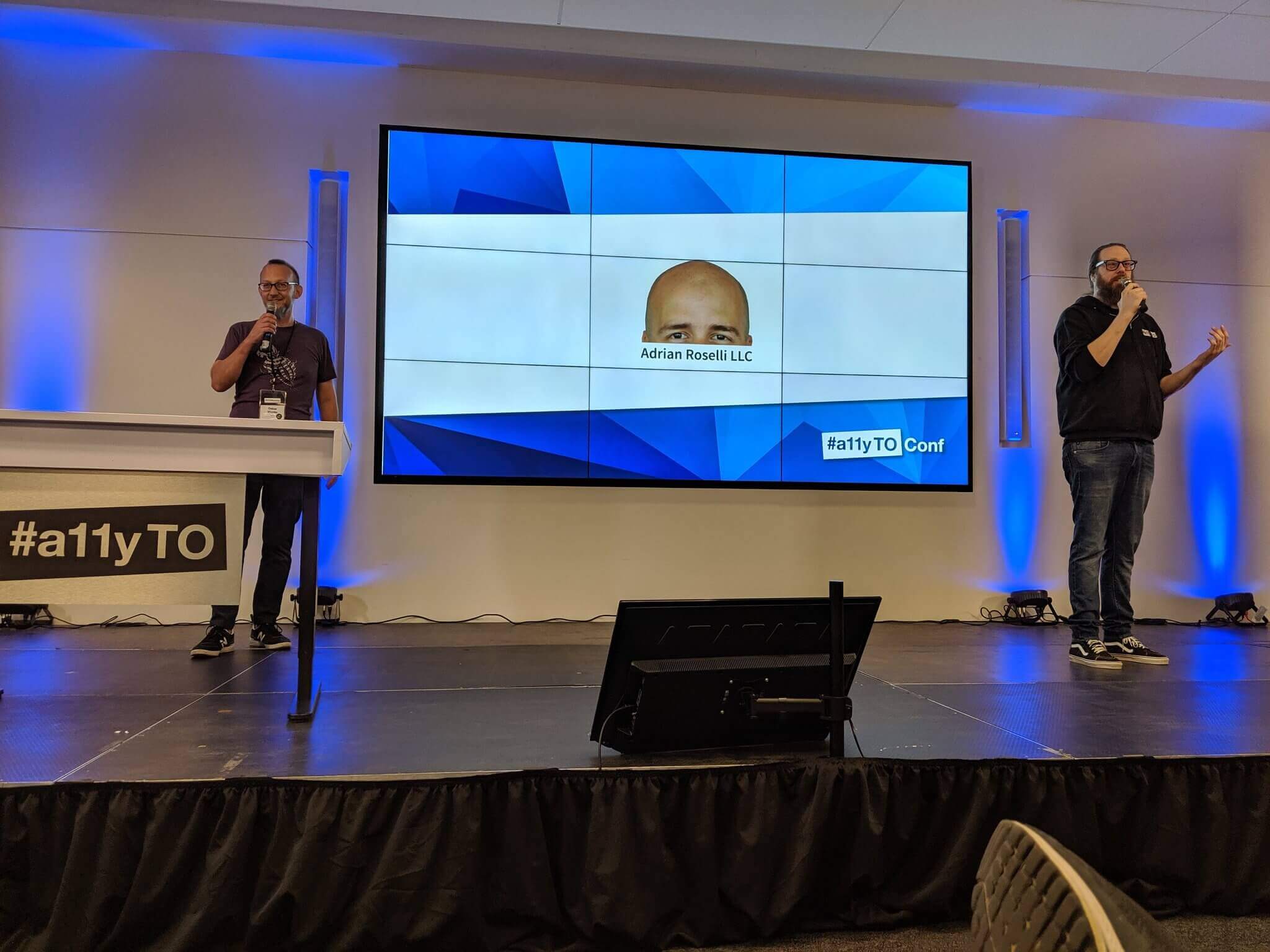 Oskar and Billy on stage in front of a large screen showing the top of my head as a sponsor.
