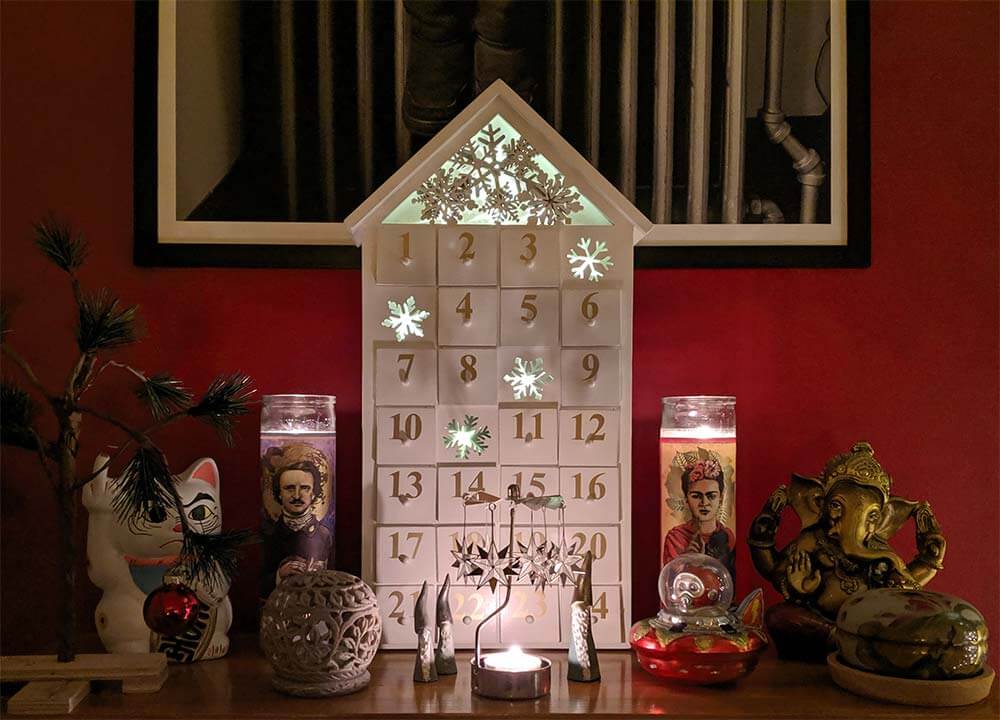 A wooden advent calendar flanked by two tall candles (one of Edgar Allen Poe, one of Frida Kahlo), a wooden Ganesh, and a lucky cat sculpture flipping off the viewer instead of waving. In the foreground is a small candle in a metal frame with a rotating metal fan top, a Charlie Brown Christmas tree, an incense burner, and three little Norwegian gnomes. All this against a red wall.
