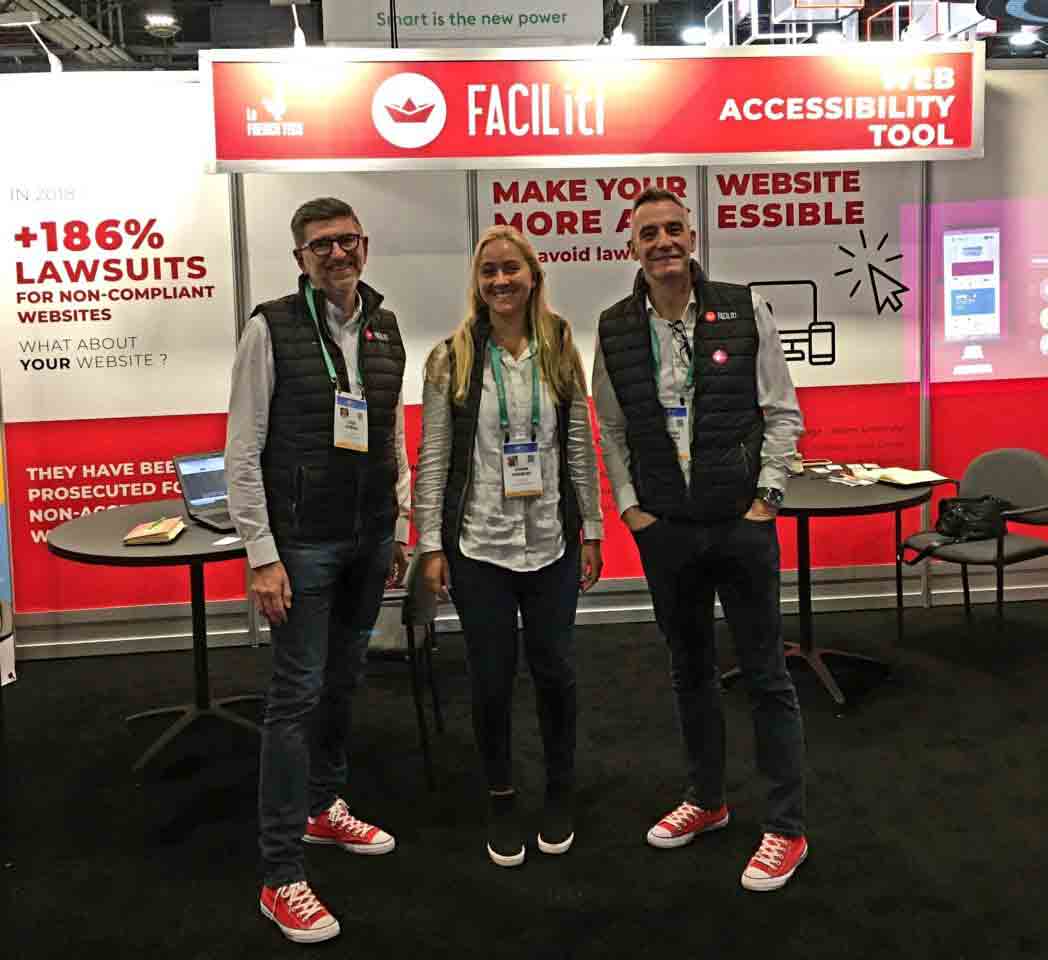 Two men and a woman wearing FACIL'iti event badges pose in front of a booth wih a prominent FACIL'iti logo.