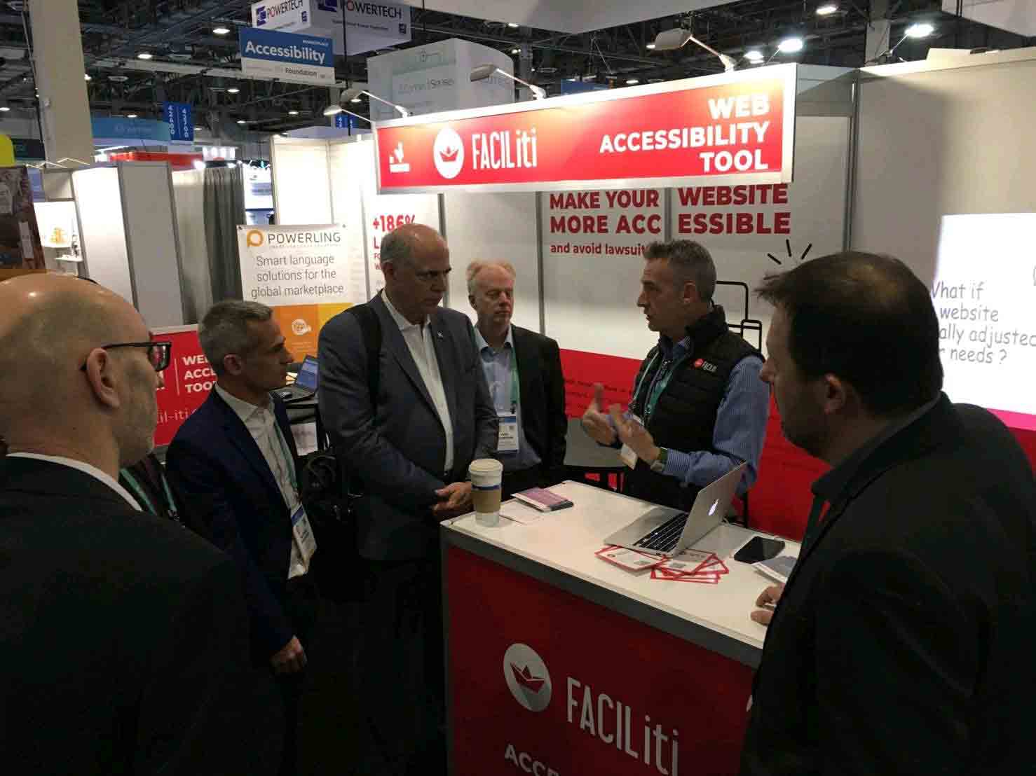 A man in a FACIL'iti vest stands at the FACIL'iti booth talking to a group of men in suits.