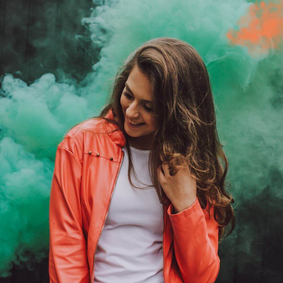 A young woman in an orange jacket is standing in a cloud of green smoke; she is stroking her long hair and looking down and away.