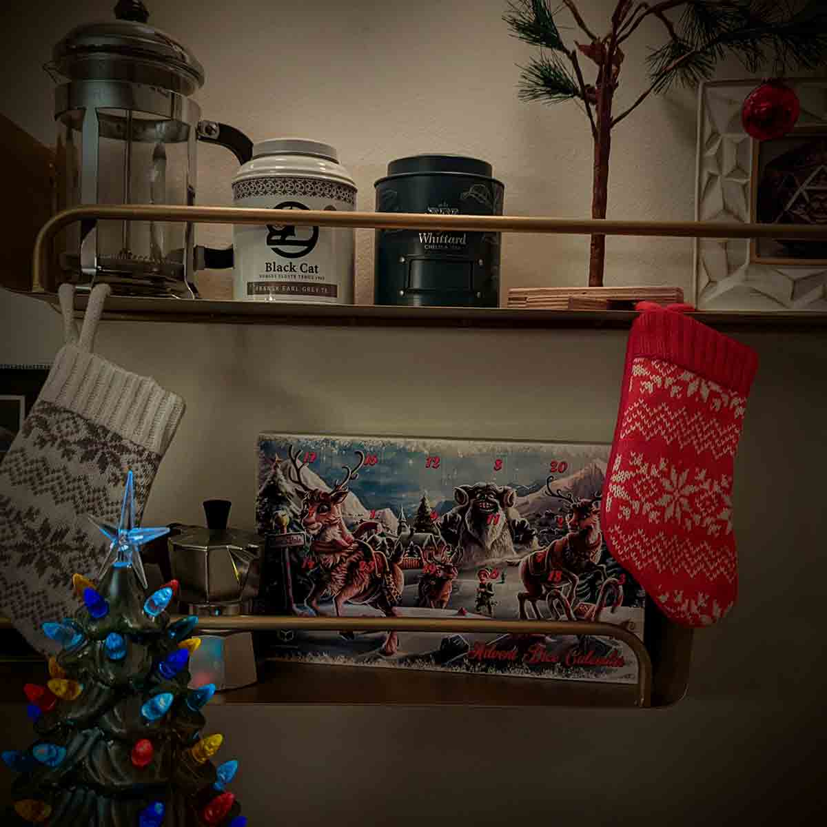 Two brass metal shelves mounted to a white wall and lit by ambient light; the top shelf has two tea tins, a French press, and a sad Charlie Brown Christmas tree along with a framed print; the second shelf has a tiny moka pot and a dice advent calendar; tiny stockings in a knit geometric snow pattern hang off the top shelf and a ceramic tree with lighted bulbs mounted to it are in the foreground.