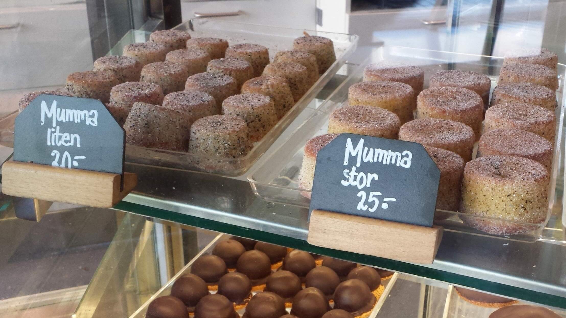 Glass case with two trays of small round cakes covered in black flecked sugar. The smaller cakes are labeled mumma liten for 20 krona, and the bigger cakes are labeled mumma stor for 25 krona. The bigger cakes are smaller than a cupcake.