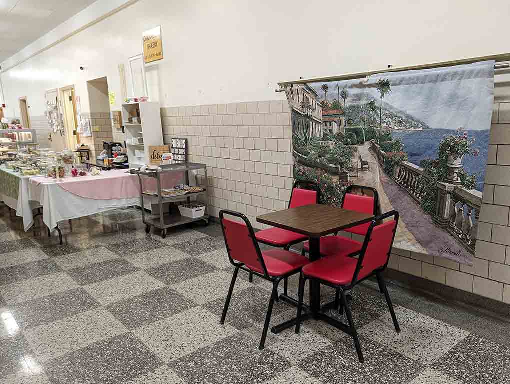 An old school hallway with institutional floors and walls; in the middle of a hallway is a beat-up table, four padded chairs, and a five by three foot fabric with an image of the Amalfi coast.