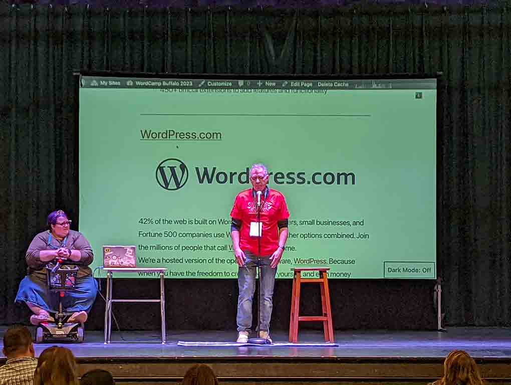 Ben Dunkle on stage in a red WCBF shirt introducing the event. Michelle Ames is behind and to the side. The screen is showing the WordPress home page.