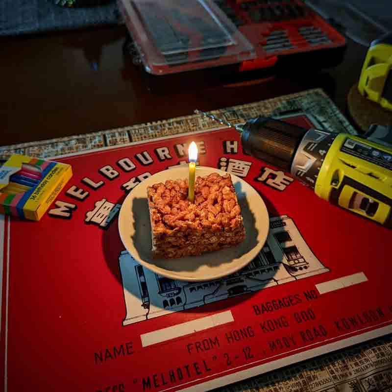 A chocolate Rice Krispie treat sits on a saucer in a darkened room, a single lit birthday candle sticking out of it. Also on the table and in shadow to the right is a drill, to the left is a pack of candles, and behind it a set of drill bits.