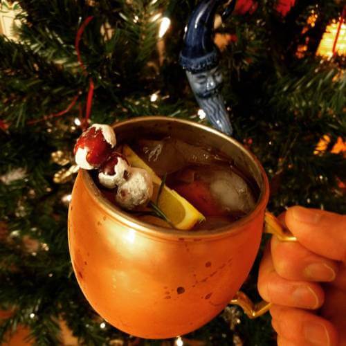 Copper mug held in front of a Christmas tree; visible in the mug is ice, sugared cranberries, an orange slice, and rosemary.