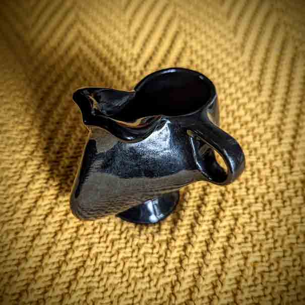 A black ceramic mug with a handle, except instead of the front of the mug following the general round shape, it extends out almost like a large human nose, and at the top of the mug where the bridge of the nose hits the rim, there are two stubby horizontal wings extending the rim.