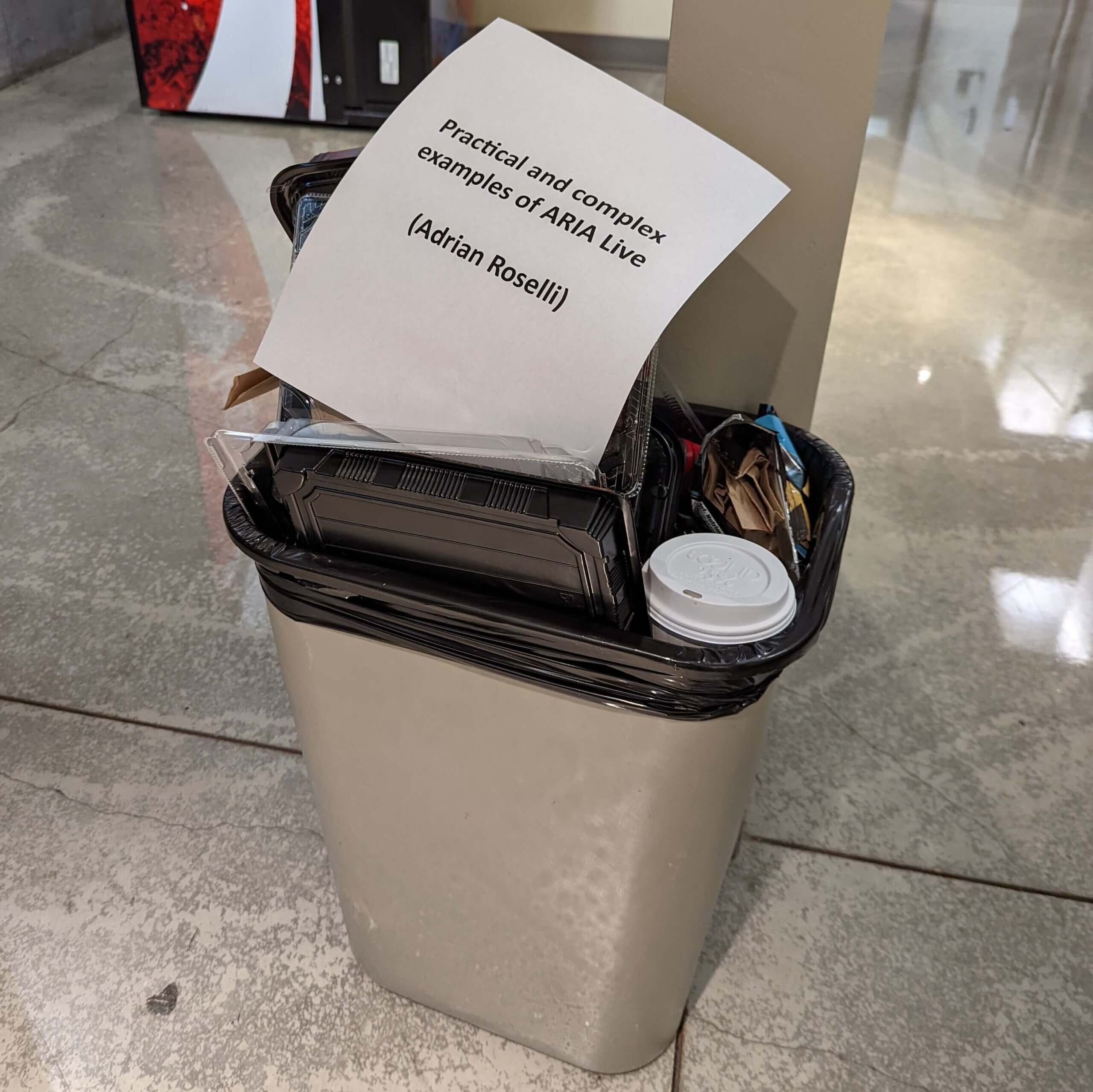 The paper sign with my name has been casually dropped onto a small rubbish bin overflowing with lunch detritus.