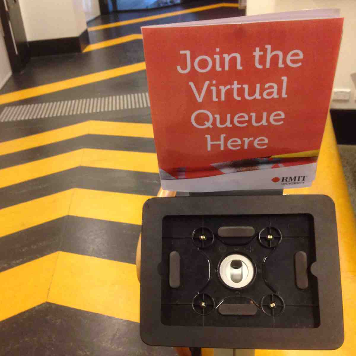 White on red sign, “Join the Virtual Queue Here” but the attached iPad mount is missing its iPad; this on a stand against a black and yellow floor striped in arrows to indicate in which direction you must move.