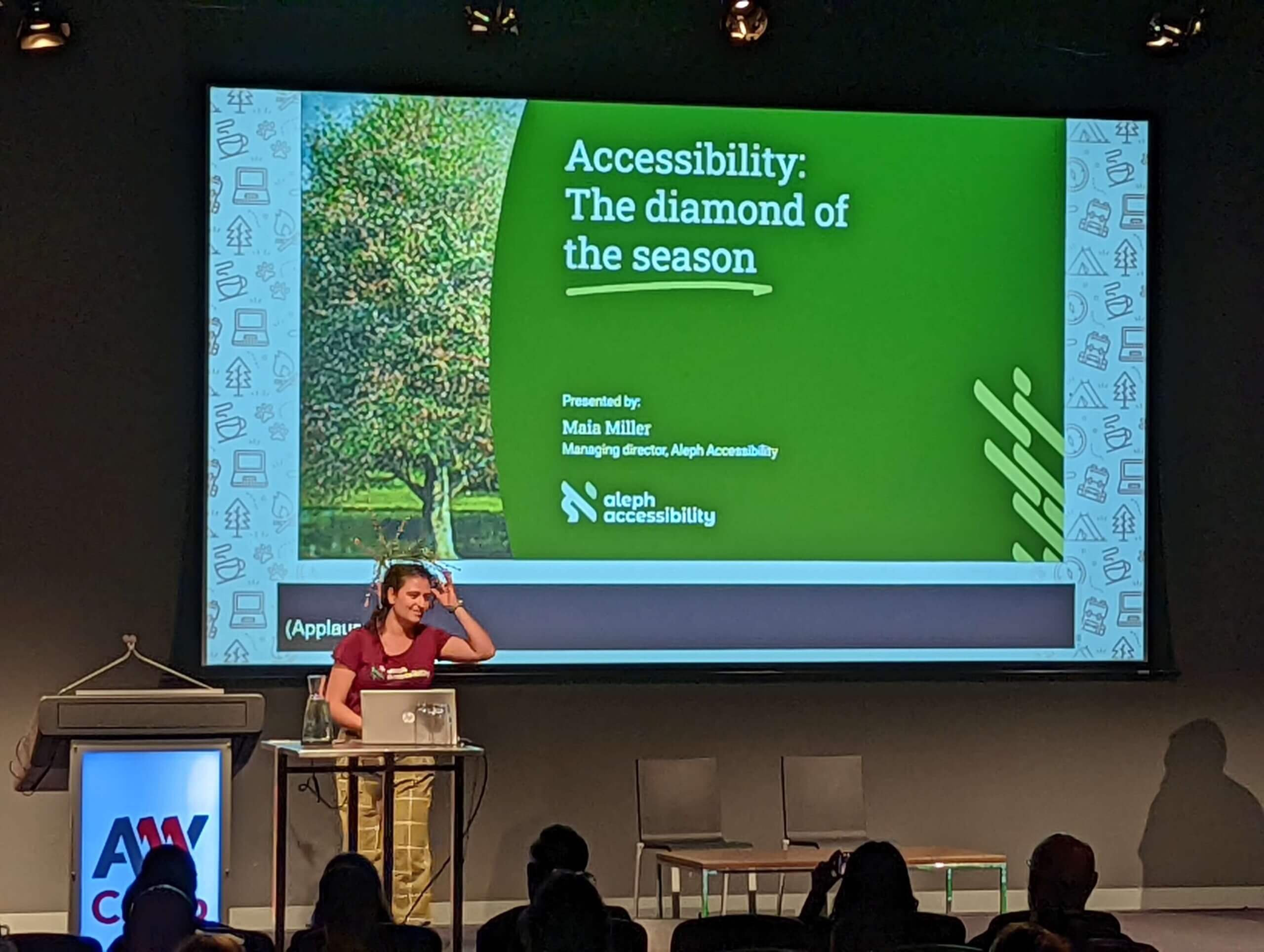 A young woman with dark hair motions to the cluster of flowers and stems sticking up from the back of her brown hair; behind her is a slide with the text, “Accessibility: The diamond of the season.”