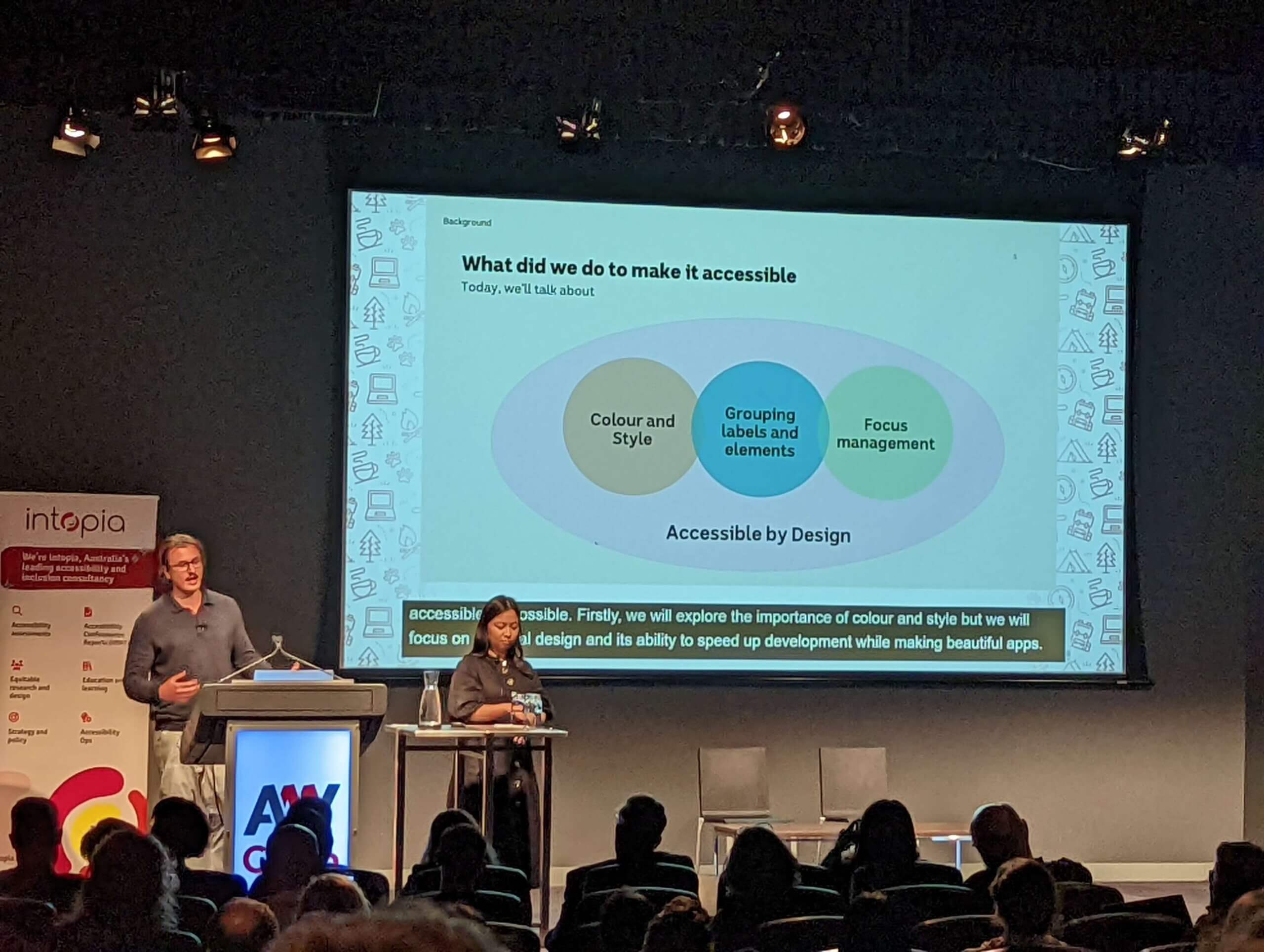 A young man and woman in front of a slide titled “What did we do to make it accessible.” It shows three circles: “Colour and style,” “Grouping labels and elements,” and “Focus management.” These are all contained in a larger oval labeled, “Accessible by Design.”