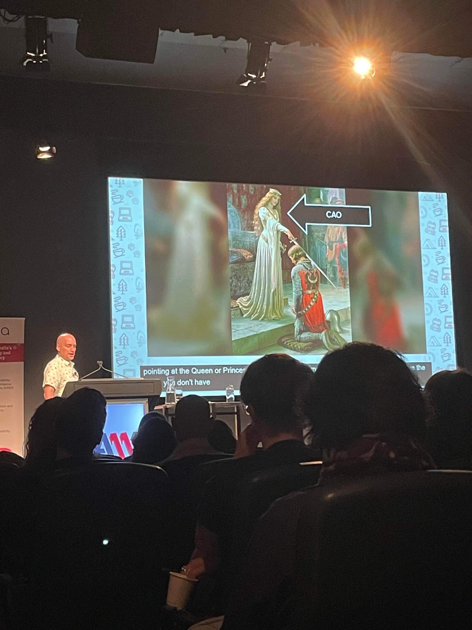 Adrian Roselli on stage next to his slides that show a painting of a queen knighting a knight, there is an arrow pointing to the queen with the label CAO.