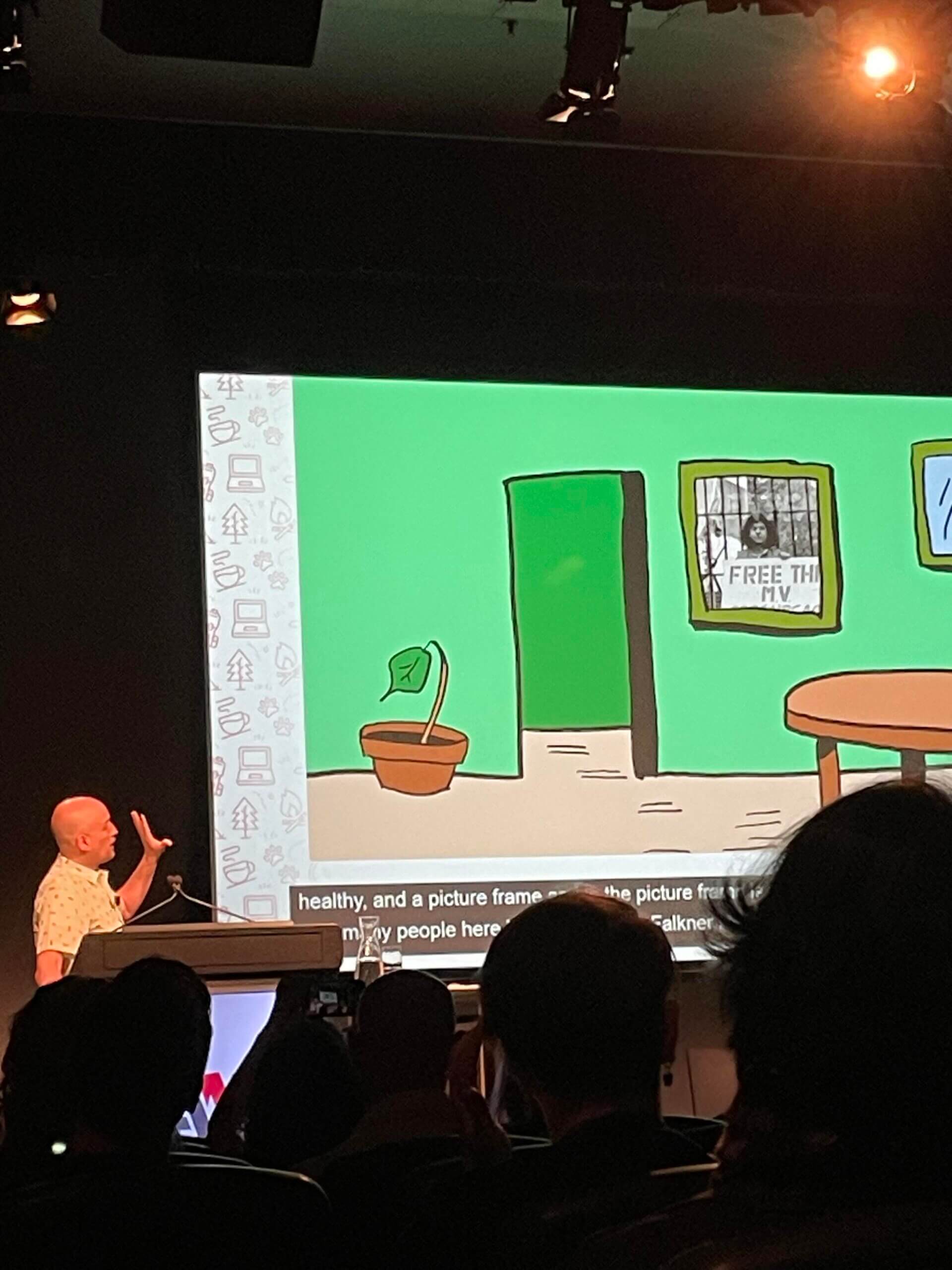 Adrian Roselli on stage at A11yCamp looking at his slides which are showing a drawing of a room with a picture frame with a photo of Steven Falkner on the wall.