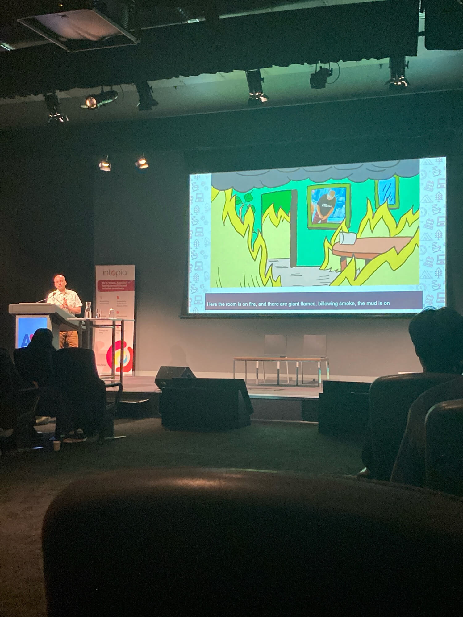 Adrian speaking on stage next to a large presentation slide. The slide shows a photo of Steve Faulkner looking downcast in a cartoon of a room full of flames.