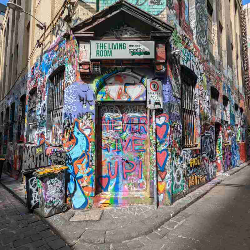 Looking straight at a corner entrance to a brick building, the ground floor of which is completely covered in rich texture of brightly colored graffiti made up of symbols, letters, and sweeps of chaos, none of which is legible except for “Never give up!” across the door.