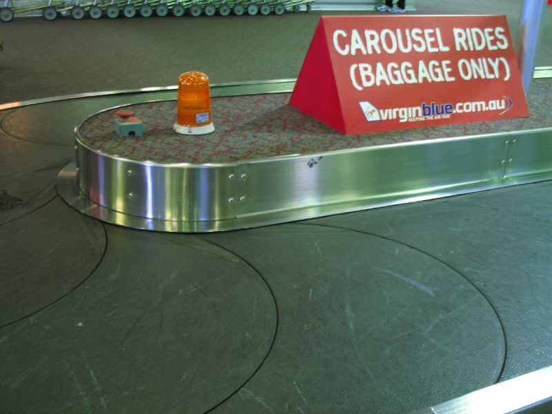 An airport luggage carousel with a sign posted above conveyor belt that reads “Carousel rides (baggage only).”