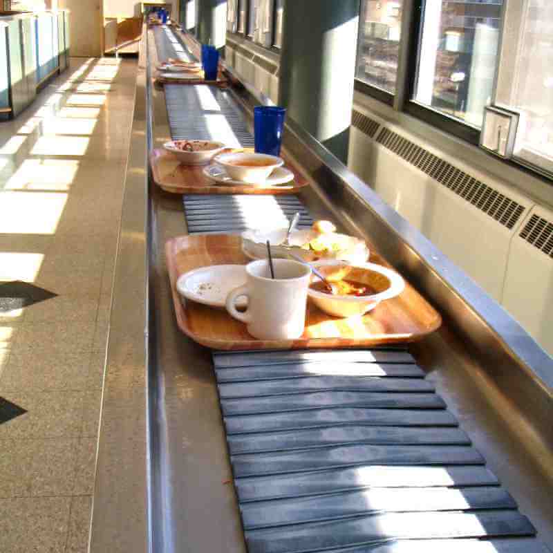 A conveyor belt in what feels like an institutional setting that has a few trays holding empty plates, bowls, and mugs, all sliding from the bottom right of the frame to a very distant hole in the wall at the top left of the frame.