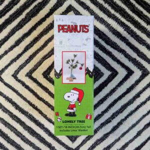 A boxed Charlie Brown lonely tree (a spindly pine stick with a single ornament bending it to the ground) on a carpet of concentric black and white diamonds.