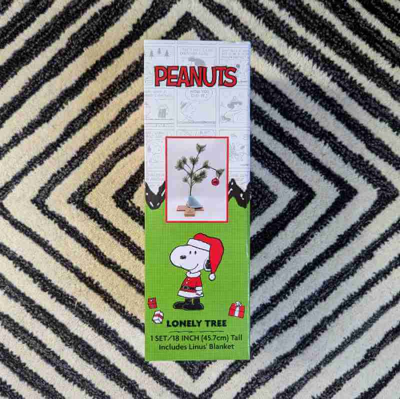 A boxed Charlie Brown lonely tree (a spindly pine stick with a single ornament bending it to the ground) on a carpet of concentric black and white diamonds.